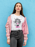 A woman stands against a bright blue textured wall wearing a white graphic T-shirt with a black and pink Medusa design and the word 'REBEL', a light pink bomber jacket, and dark gray jeans.
